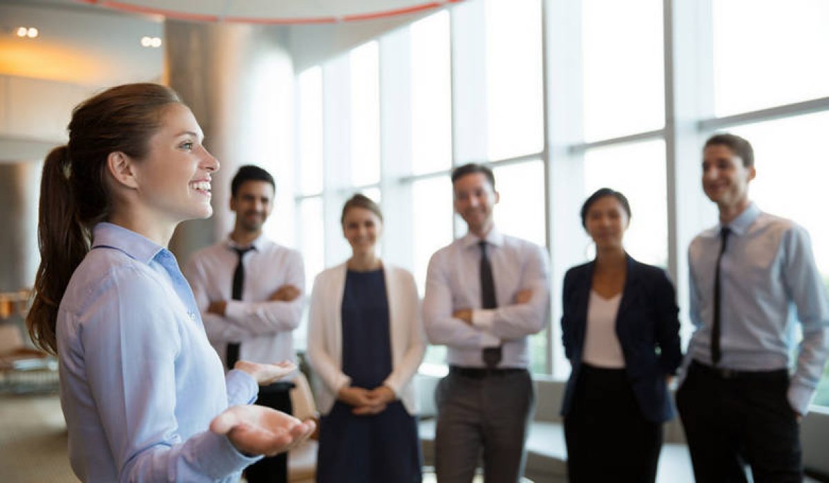 Portrait of successful female executive manager with open hands gesture standing in front of her team in office hall and announce good news